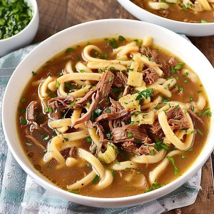 Bowl of slow-cooked beef noodle soup garnished with green onions and herbs.
